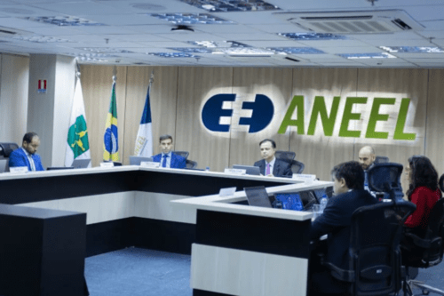 Uma sala de reuniões formal da ANEEL (Agência Nacional de Energia Elétrica). Ao fundo, em uma parede de madeira clara, destaca-se o logotipo azul e verde da agência. Seis pessoas estão sentadas ao redor de uma bancada em formato de "U" na cor preta e branca, utilizando notebooks e papéis. Três bandeiras oficiais estão posicionadas à esquerda. O ambiente é bem iluminado, com teto de placas de escritório e ar-condicionado central.