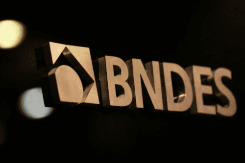 Fotografia em close do logotipo metálico do BNDES (Banco Nacional de Desenvolvimento Econômico e Social). As letras são tridimensionais, com acabamento em metal escovado dourado ou bronze, destacando-se contra um fundo escuro e desfocado com pontos de luz suaves (efeito bokeh).