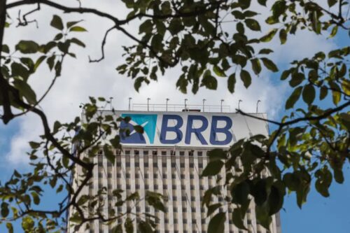Uma fotografia em ângulo baixo (contra-plongée) mostra o topo de um edifício comercial imponente onde se lê o letreiro do BRB (Banco de Brasília).