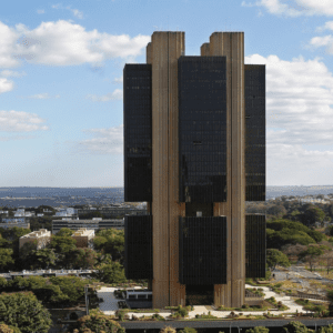 Vista aérea do edifício-sede do Banco Central do Brasil em Brasília, uma torre de arquitetura brutalista cercada por vegetação e céu com nuvens.