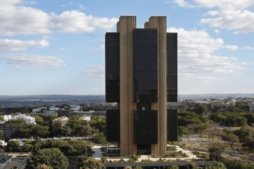 Vista aérea do edifício-sede do Banco Central do Brasil em Brasília, uma torre de arquitetura brutalista cercada por vegetação e céu com nuvens.