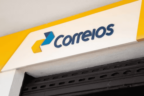 Close da fachada de uma agência dos Correios. A imagem destaca o logotipo moderno da empresa em azul e amarelo sobre um fundo branco, posicionado acima de uma porta de enrolar preta entreaberta.