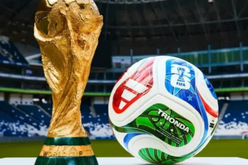 Troféu da Copa do Mundo da FIFA em destaque ao lado da bola oficial do torneio de 2026, posicionados no centro de um estádio moderno com arquibancadas ao fundo.