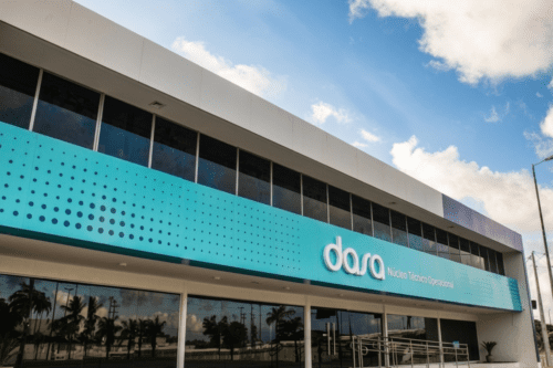 Fachada moderna do Núcleo Técnico Operacional da Dasa. O prédio possui dois andares, com janelas de vidro escuro e um grande painel azul claro com estampa de retículas. O logotipo branco da 'dasa' está centralizado no painel. O céu está azul com nuvens brancas e há palmeiras refletidas nos vidros do térreo.