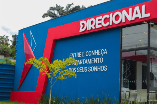 Fachada de um estande de vendas da construtora Direcional. O prédio possui paredes externas em azul vibrante com detalhes estruturais em vermelho que formam uma moldura geométrica. No topo, o logotipo da "Direcional" em letras brancas sobre um fundo vermelho. Na parede azul, lê-se a frase: "Entre e conheça o apartamento dos seus sonhos". À direita, uma grande vitrine de vidro permite ver o interior iluminado com mesas e cadeiras brancas. No primeiro plano, há pequenas plantas ornamentais e gramado.