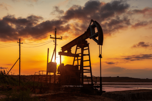 Imagem de uma torre de extração de petróleo ao entardecer, com o céu cheio de nuvens e tons quentes do pôr do sol, destacando-se como símbolo de energia e petróleo.