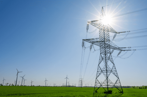 Vista de torres de transmissão de energia elétrica em um campo com parques eólicos ao fundo sob céu claro e ensolarado.