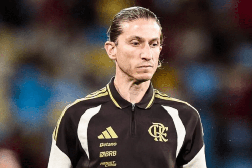 Fotografia em plano médio de Filipe Luís, ex-jogador e atual treinador de futebol. Ele aparece com expressão séria, olhando levemente para baixo. Seu cabelo castanho está preso em um coque justo. Ele veste um uniforme de treino do Flamengo na cor preta, com detalhes em dourado e branco nos ombros, exibindo os logotipos da Adidas e o escudo do clube (CRF) no peito, além de patrocinadores como Betano e BRB. O fundo está desfocado, sugerindo o ambiente de um estádio de futebol.