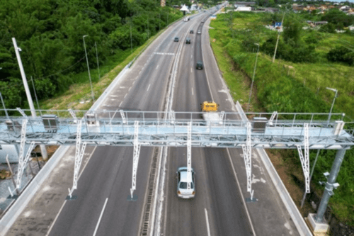 Sistema de pedágio eletrônico free flow instalado sobre rodovia, com veículos passando sem parar em pista cercada por vegetação.