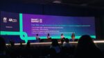 Foto tirada da plateia durante o painel "Copa 2026 e Copa Feminina: O Futuro do Futebol e o Impacto do Esporte na Economia" no evento SMART Summit 26. No palco, quatro pessoas estão sentadas em poltronas participando de uma discussão, com um telão ao fundo apresentando os nomes e cargos dos participantes: Marina Fuzeti Fagali, Guga Menga, Pretinha e a host Bruna Allemann.