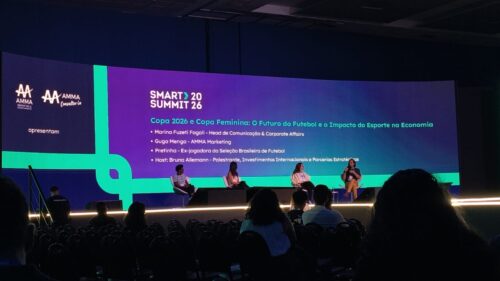 Foto tirada da plateia durante o painel "Copa 2026 e Copa Feminina: O Futuro do Futebol e o Impacto do Esporte na Economia" no evento SMART Summit 26. No palco, quatro pessoas estão sentadas em poltronas participando de uma discussão, com um telão ao fundo apresentando os nomes e cargos dos participantes: Marina Fuzeti Fagali, Guga Menga, Pretinha e a host Bruna Allemann.