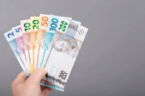Mão segurando um leque de notas do Real brasileiro (de 2 a 200 reais), representando o poder de compra e o impacto da inflação na economia, contra um fundo cinza neutro.
