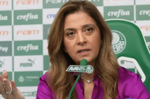 Retrato em plano médio da empresária Leila Pereira, presidente do Palmeiras, falando durante uma coletiva de imprensa. Ela veste uma camisa de cetim magenta e gesticula com a mão direita. Ao fundo, um painel com logotipos de patrocinadores e o escudo do clube, além de um microfone com a marca "Avanti" posicionado à frente.