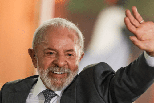 Close-up do presidente Luiz Inácio Lula da Silva sorrindo e acenando com a mão esquerda levantada. Ele possui cabelos e barba grisalhos, usa terno cinza, camisa branca e gravata azul com padrões geométricos. O fundo está em tons de laranja e bege, suavemente desfocado.