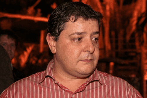 Fotografia em close de um homem de meia-idade com cabelos escuros e levemente ondulados, vestindo uma camisa social de botões com listras verticais brancas e vermelhas. Ele tem uma expressão séria e olha para o lado. O fundo está desfocado, apresentando uma iluminação quente em tons de laranja e sombras, sugerindo um ambiente externo noturno com vegetação.