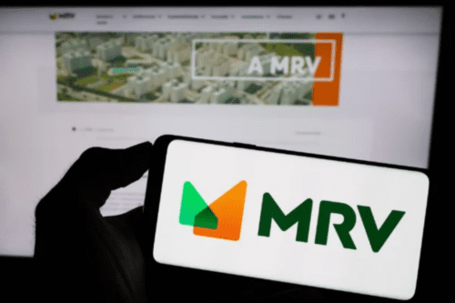 Uma mão segura um smartphone em primeiro plano exibindo o logotipo colorido da MRV (composto por formas geométricas em verde e laranja). Ao fundo, em desfoque, vê-se a tela de um computador mostrando o site oficial da empresa com imagens de empreendimentos imobiliários.