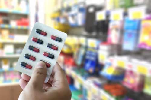 Primeiro plano de uma mão segurando uma cartela de medicamentos (blister) com 10 cápsulas nas cores vermelho e preto. O fundo da imagem está desfocado, mostrando prateleiras coloridas de um estabelecimento comercial, como um supermercado ou farmácia, com diversos produtos e etiquetas de preço amarelas.