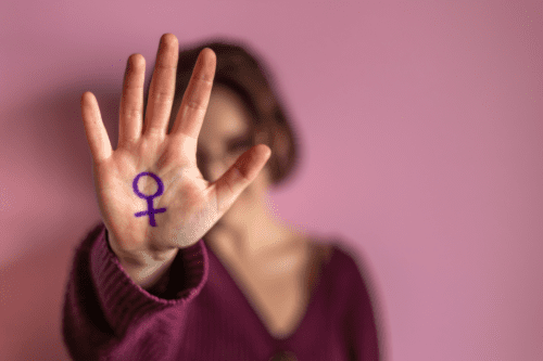 Mulher segurando a mão com símbolo feminino pintado na palma contra fundo rosa. Representação do empoderamento feminino e luta pelos direitos das mulheres.