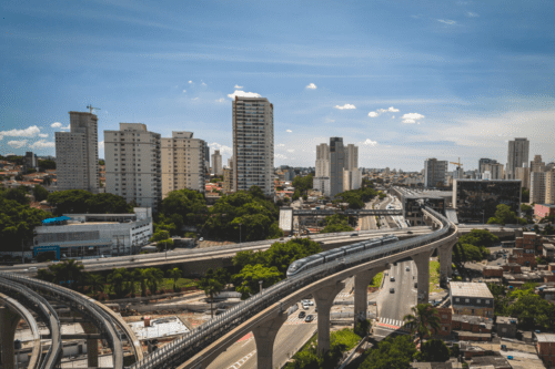 Uma vista aérea panorâmica de uma metrópole moderna, destacando um monotrilho de design futurista e prateado que percorre trilhos elevados em primeiro plano.