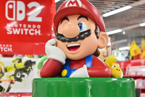 close-up de um display de loja mostrando um busto grande de plástico de Mario, do Mario Kart 8 Deluxe. Ele está em um Cano Verde, apoiando o queixo na mão com um sorriso e uma expressão pensativa. Ao lado dele estão pelúcias de Luma e Yoshi Amarelo. O fundo é uma seção de jogos de um varejista, com um grande display vermelho da Nintendo Switch visível.