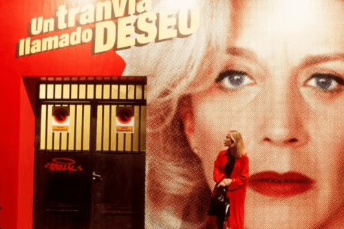 Gal Costa vestindo um sobretudo vermelho e segurando uma bolsa preta está parada em frente a uma parede vermelha com uma grande imagem impressa do rosto de uma mulher. Acima da imagem, lê-se em letras grandes: "Un tranvía llamado DESEO". À esquerda, há um portão de ferro escuro com dois sinais de proibido estacionar.