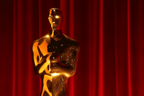 Estatueta dourada do Oscar em destaque, com acabamento brilhante, posicionada à frente de uma cortina de veludo vermelho.
