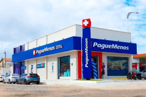 Fotografia da fachada de uma unidade da Farmácia Pague Menos em uma esquina ensolarada. O prédio é moderno, de cor bege, com uma grande testeira azul onde se lê o nome da marca e o logotipo de uma cruz branca sobre fundo vermelho. Há um totem vertical azul alto também com a marca "Pague Menos". Carros estão estacionados na lateral esquerda e o céu ao fundo está claro com poucas nuvens.