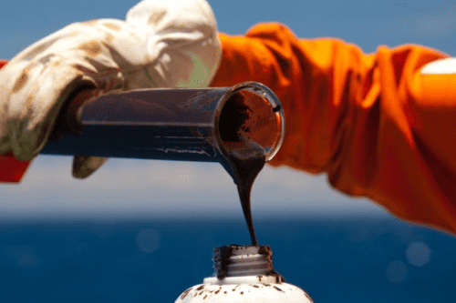 Foto em close de uma pessoa com luvas e uniforme laranja despejando petróleo bruto de um tubo de ensaio de vidro em um frasco branco, com o mar azul ao fundo.