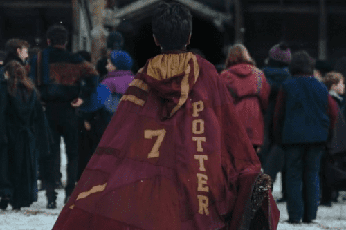 Vista traseira de um jovem bruxo usando uma capa de Quadribol da Grifinória, personalizada com o nome "POTTER" e o número "7" costurados em tons de dourado sobre o tecido carmesim. Ele está de frente para uma multidão em um ambiente invernal.