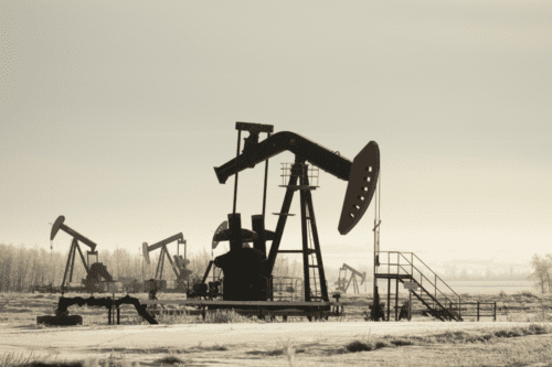 Imagem de várias torres de perfuração de petróleo em um campo petrolífero sob céu nublado, destacando-se pela silhueta escura contra o clima frio e desolado.