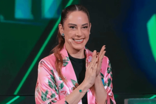 A apresentadora Silvia Abravanel sorri e bate palmas em um estúdio de televisão. Ela veste um blazer de cetim rosa com estampas florais verdes sobre uma blusa preta, e usa um microfone auricular.