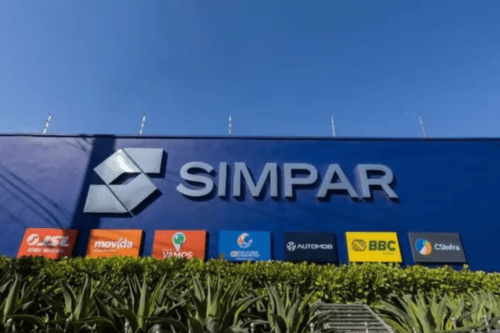 Imagem da fachada da sede da empresa Simpar, com o logo da companhia e várias marcas de parceiros e associados visíveis na frente, sob céu azul claro.