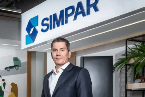 Retrato de executivo da Simpar, vestindo paletó azul-marinho e camisa social branca, posicionado à frente do logotipo da empresa em um ambiente corporativo moderno.