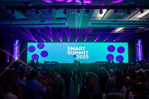 Vista em ângulo aberto de um auditório lotado durante o evento 'Smart Summit 2026'. Ao fundo, um palco amplo é dominado por uma tela digital gigante de cor azul-turquesa vibrante, onde se lê 'SMART SUMMIT 2026' com um ícone de seta ascendente. A tela é ladeada por formas geométricas de hexágonos estilizados. O palco é iluminado por vários feixes de luz roxa intensa que se projetam para o teto a partir da base da tela. Nas laterais, painéis verticais exibem o nome do evento. O público, visto de costas e em silhueta, ocupa as poltronas da plateia, criando um contraste entre a iluminação cênica do palco e a penumbra do auditório.