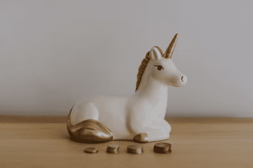 Estatueta de um unicórnio branco e dourado sobre uma mesa de madeira, acompanhada de pequenas pilhas de moedas, representando o conceito financeiro de empresas "unicórnio".
