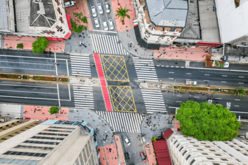 Vista aérea em ângulo zenital do cruzamento das avenidas Ipiranga e São João, no centro de São Paulo. A imagem mostra o fluxo de veículos e pedestres em torno de um cruzamento com sinalização de solo, incluindo faixas de pedestres e áreas de bloqueio (cruzamento amarelo), com edifícios antigos nas esquinas e uma grande árvore à direita, local cotado para a implementação de painéis de LED do projeto 'Boulevard São Paulo'.