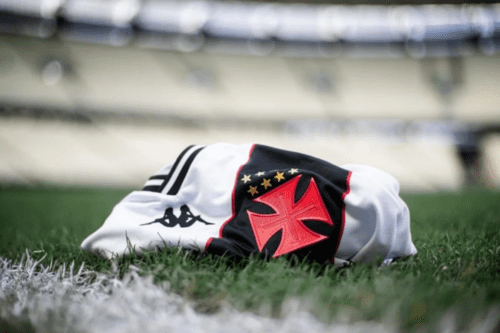 Camisa oficial do Vasco da Gama, branca com a faixa diagonal preta, dobrada sobre o gramado verde de um estádio de futebol. Em destaque, a Cruz de Malta vermelha e as estrelas douradas do escudo, ao lado do logotipo da Kappa. O fundo mostra as arquibancadas do estádio desfocadas.