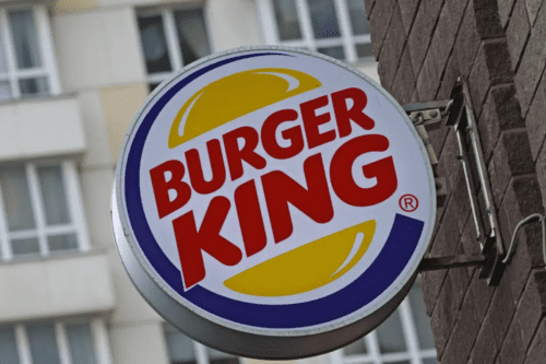 Logotipo circular do Burger King em uma placa externa fixada em uma parede de tijolos marrons. O logo apresenta o nome da marca em vermelho, entre duas metades de um pão amarelo, circundado por uma linha azul. Ao fundo, vê-se a fachada desfocada de um edifício residencial.