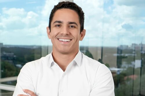 Lucas Feitosa, diretor da Avenue, sorridente de camisa branca em escritório moderno, com vista urbana ao fundo, em entrevista sobre investimentos e dólar.