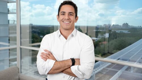 Lucas Feitosa, diretor da Avenue, sorridente de camisa branca em escritório moderno, com vista urbana ao fundo, em entrevista sobre investimentos e dólar.