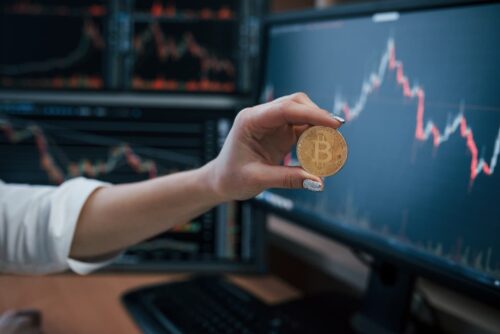 Mão segura moeda de bitcoin em frente a telas com gráficos de mercado financeiro em queda e alta.