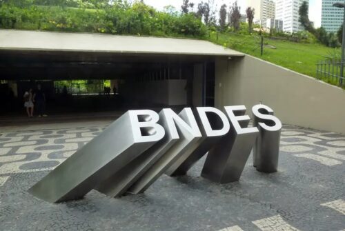 Escultura com a sigla BNDES na entrada de prédio, com área verde acima e passagem subterrânea ao fundo.