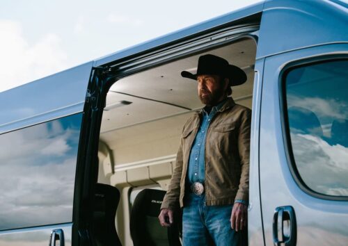 Chuck Norris em pé na porta de uma van, olhando ao longe em cenário aberto.