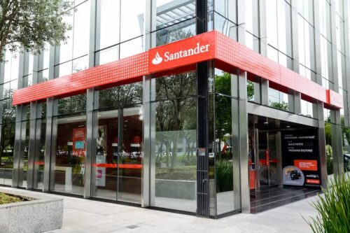 Agência do Santander com fachada moderna em vidro e logotipo vermelho em destaque na entrada do banco.