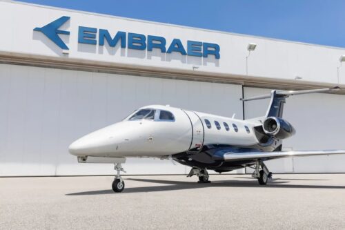 Jato executivo estacionado em frente a hangar com logotipo da Embraer, em área industrial de aeroporto, sob céu azul claro.