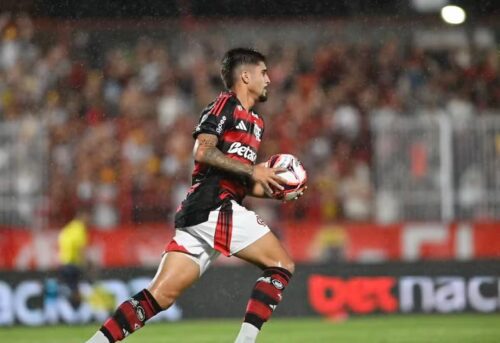 Jogador do Flamengo em campo com a bola durante partida, com torcida ao fundo em noite de jogo.