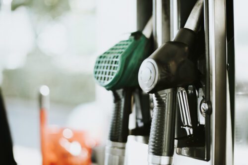 Uma foto em close de duas bombas de combustível em um posto de gasolina. A bomba em primeiro plano é preta, enquanto a que está logo atrás é verde, ambas com mangueiras cinzas. O fundo está desfocado, mostrando tons de verde e um pouco de laranja à esquerda. A iluminação é natural e suave, com foco nítido nos bicos das bombas.
