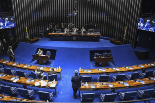 Vista ampla do plenário do Senado Federal no Brasil, mostrando as bancadas azuis, parlamentares e a mesa diretora em sessão.