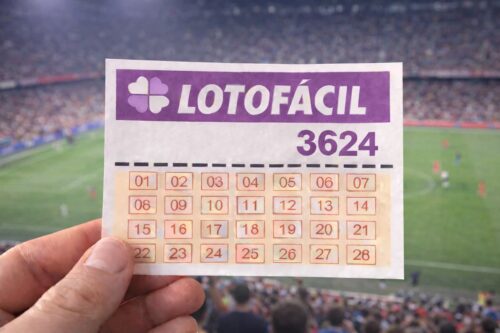 Bilhete Lotofácil 3624 segurado em estádio lotado durante partida de futebol ao fundo desfocado.