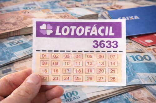 Bilhete da Lotofácil 3633 segurado por mão sobre notas de real, com cartões e caderneta da Caixa ao fundo.